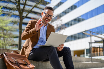 Happy satisfied businessman sitting on steps and showing OK sign over laptop during online meeting