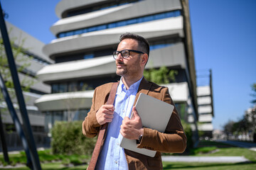 Serious entrepreneur with laptop looking away and standing against modern office building in city