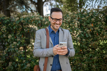 Handsome professional in eyeglasses with bag messaging online over mobile phone against plants