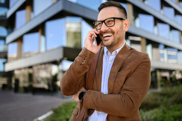 Young businessman smiling and talking over phone call while standing against modern building in city