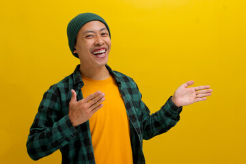 Excited young Asian man, dressed in a beanie hat and casual shirt, extends his arms to the side towards an empty space, inviting someone to join him while standing against a yellow background