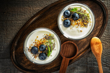 Two glass jars of yogurt with blueberries, chia seeds, osmanthus flowers and pea shoots on a wooden oval tray, with two wooden spoons. View from above.