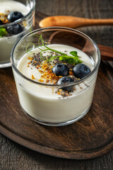 Homemade yogurt on a wooden tray with a wooden spoon on a textured gray wooden background, garnished with blueberries, chia seeds, dried osmanthus flowers and pea sprouts. Side and top view.