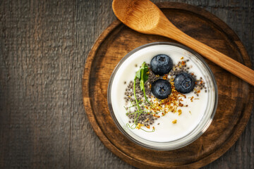 Homemade yogurt in a glass cup decorated with blueberries, chia seeds, osmanthus flowers and pea sprouts on a textured wooden plate with a wooden spoon, on a gray wooden background. View from above.