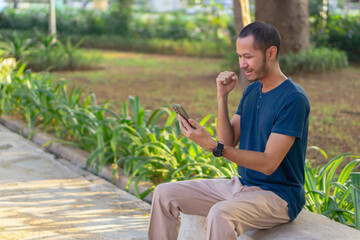 Young asian men with blue shirt smiling and laughing while playing smartphone on the park outdoor