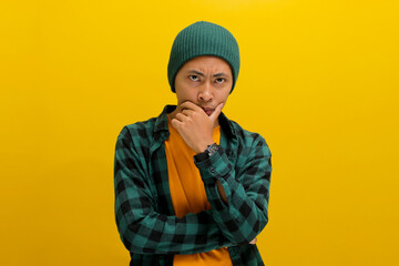 Pensive young Asian man, wearing a beanie hat and casual shirt, places his hand on his chin, looking at the camera with a confused expression, deeply lost in thought and contemplating the future