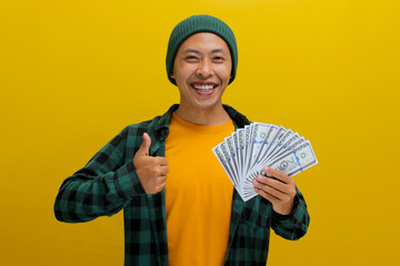 Enthusiastic Asian man in a beanie and casual clothes flashes a thumbs-up and waves a wad of cash....