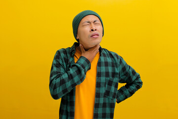 Young Asian man in beanie hat and casual shirt is experiencing a sore throat, possibly due to irritation or an infection, Isolated on yellow background. Healthcare and medical concept.