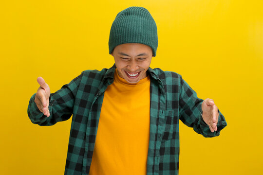 A young Asian man, dressed in casual clothes and wearing a beanie hat, is using hand gestures to indicate a big or large size while looking down, conveying the concept of measurement or size