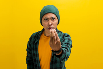 Annoyed young Asian man, dressed in a beanie hat and casual shirt, makes the Italian hand gesture, pinching his fingers together, while standing against a yellow background
