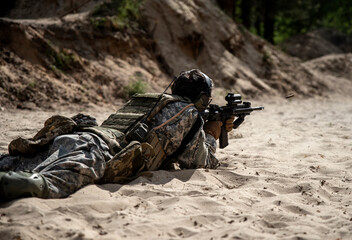 A soldier in camouflage shoots from a machine gun while lying on the sand