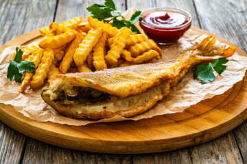 Fried sea bass served on paper with French fries and ketchup on board on wooden table 