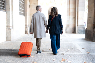 Smiling middle-aged Caucasian tourist couple walking hand-in-hand carefree with their luggage...