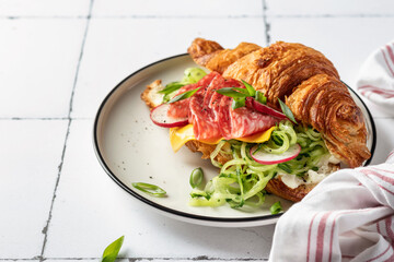 Croissant sandwich with salami sausage, cheese and cucumber in a plate for tasty breakfast on white...