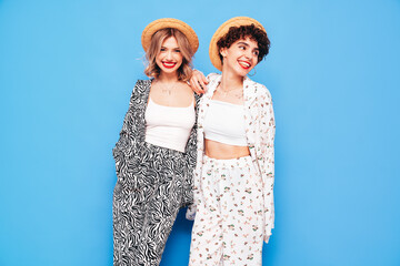 Two young beautiful smiling brunette hipster female in trendy summer costume clothes. Sexy women posing near blue wall in studio. Positive models having fun. Cheerful and happy. In hat, red lips