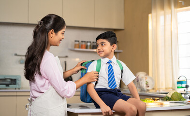 Caring Indian mother preparing kid for school by placing lunch box and water in bad - concept of responsibility, parental caring and back to school