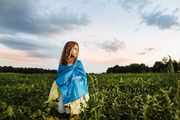 A beautiful girl in a soybean field poses with a yellow-blue flag of Ukraine at sunset. The child looks into the distance against the background of a blue cloudy sky and green nature.