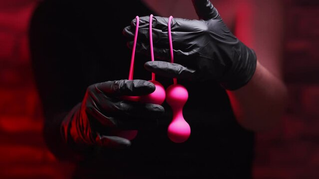 Woman's hand in black latex gloves and a black dress holds pink Kegel balls and Ben Wa balls on a red background. Vaginal balls in a woman's hand. Sex toys Geisha Balls, Products for intimate stores
