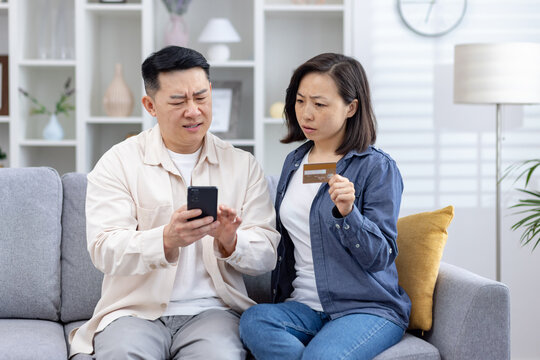 Asian couple looks distressed as they uncover unauthorized charges on their credit card phone, man confusion while the woman, holding the card, seems anxious. stress, frustration of financial fraud. - Powered by Adobe