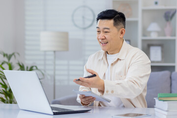 Asian man interacts with a webinar on his laptop at home, expressing happiness and engagement. This...