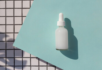 Cosmetic serum glass bottle on the blue background with white ceramic tile and tropical leaf shadow.