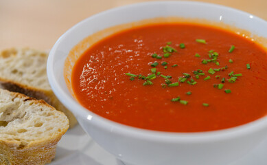 A white bowl of tomato and basil soup with two slices of baguette bread on a plate.
