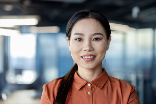 Confident Asian businesswoman in a bright office environment. She stands elegantly dressed in a burnt-orange shirt, signaling professionalism and approachability.