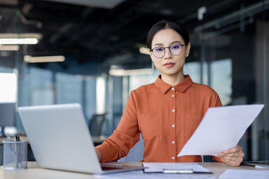 A professional Asian businesswoman examines paperwork with a serious expression while sitting at her office desk. Perfect scene of focused work.