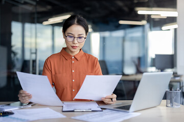 A serious young professional woman in a stylish orange shirt reviews paperwork intently, surrounded by laptops in a well-lit office setting. Her focus and determination are evident.
