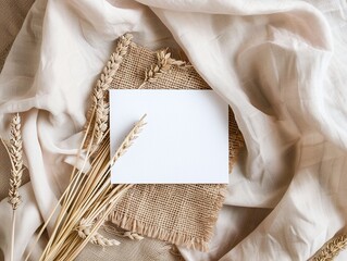 Blank white card on a beige silk fabric with golden wheat ears. Flat lay composition with copy space. Harvest and invitation design concept for greeting card and print.