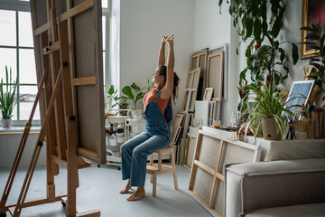 Relaxed woman artist stretching sitting on chair sighing deeply looking out window. Calm full of...