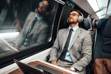 Tired handsome businessman relaxing while traveling with high-speed train or metro. He is using laptop computer and wireless headphones for entertainment and music listening. Modern travel concept.