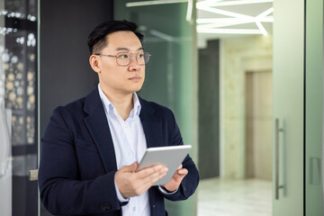 An Asian businessman focuses intently on a tablet while standing in a sleek, modern office corridor, symbolizing contemporary business practices.