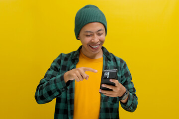 An excited young Asian man, dressed in a beanie hat and casual shirt, points at his mobile phone while standing against a yellow background