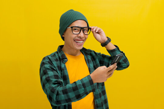 Asian man, dressed in a beanie hat and casual shirt, adjusts his eyeglasses and peeks above them, smiling as he looks at the camera with a mobile phone in hand while standing against yellow background