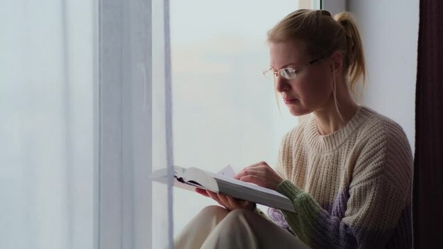 Adult woman in casual clothing reads a book while sitting on the windowsill in the city apartment. Female's intellectual leisure in a cozy home environment on a cold winter day.