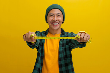 Cheerful Asian man in a beanie and casual clothes stretches a yellow measuring tape in front of...