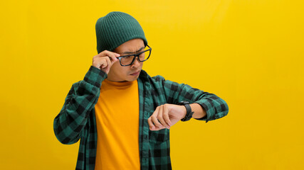 Asian man in a beanie and casual shirt checks the time on his wristwatch, isolated on a yellow...