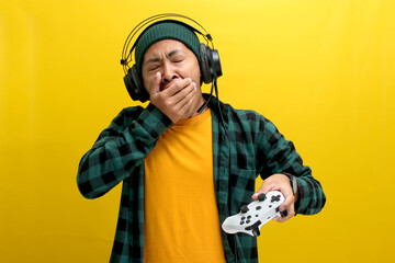 A bored Asian man, wearing headphones, a beanie hat, and a casual shirt, yawns and looks tired...