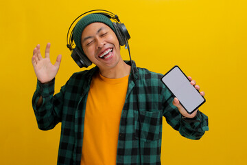 Asian man in a beanie and casual clothes listens to music on his headphones, phone held in hand....