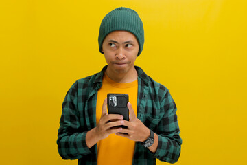 Dreamy young Asian man, wearing a beanie hat and casual shirt, holds a mobile phone with a dreamy...