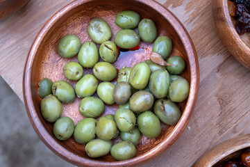 A bowl of green olives sits on a wooden table. The olives are spread out and appear to be ripe