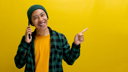 Excited young Asian man, dressed in a beanie hat and casual shirt, engages in a phone call while...