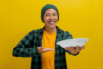 Excited young Asian man, dressed in a beanie hat and casual clothes, displays an empty white plate...