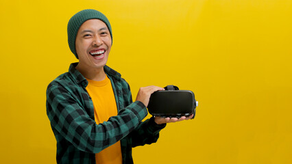 Happy Asian man holds a VR headset towards the camera, ready to explore virtual reality. Isolated on a yellow background.