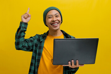 Happy young Asian man, dressed in a beanie hat and casual shirt, stands against a yellow...