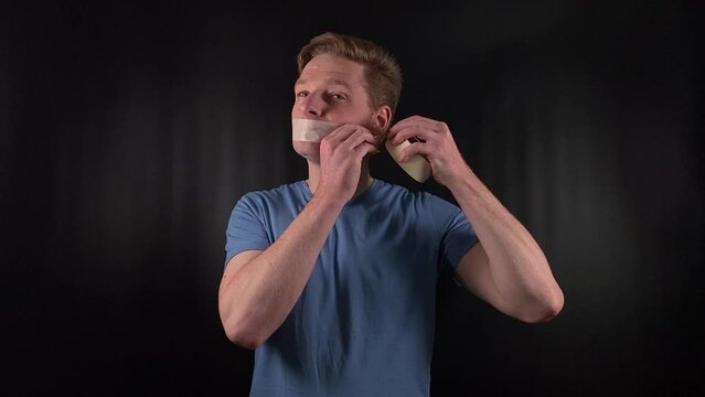A portrait of a handsome young white man taping over his mouth with adhesive tape, depicting the concept of censorship, suppression of free speech and thought on a black background. An act of protest