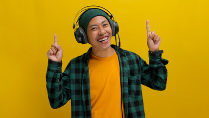 Excited Asian man in a beanie and casual clothes, wearing headphones and enjoying music or a podcast, points excitedly towards empty copy space with a happy expression. Isolated on a yellow background