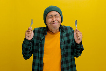 The ravenous young Asian man, sporting a beanie hat and casual shirt, closes his eyes in sheer anticipation, vividly imagining delectable food, clutching a spoon and fork against the yellow backdrop