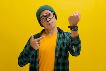 Frowning Asian man in a beanie and casual shirt impatiently taps his wristwatch, glancing at the...